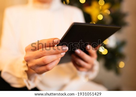 Woman in white reading interesting e-book reader while sitting near Christmas tree at home. Cozy winter pic of lady relaxing alone with novel.