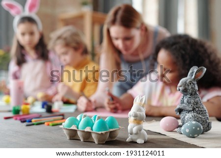 Grey and white toy rabbits and group of painted Easter eggs in egg-box standing on wooden table with kids drawing pictures on background