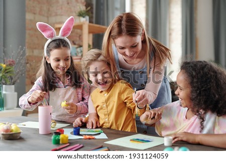 Young cheerful woman bending over table with group of happy kids while helping them with painting picture of Easter egg before holiday