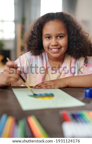 Cheerful mixed-race little girl painting large Easter egg on blank paper while sitting by wooden table and looking at you with toothy smile