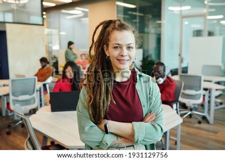Young smiling businesswoman or designer in casualwear crossing her arms by chest while standing in front of camera in working environment Royalty-Free Stock Photo #1931129756