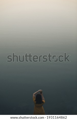 young woman in yellow dress taking picture of grey lake. evening dusk time. dark mood. copy space