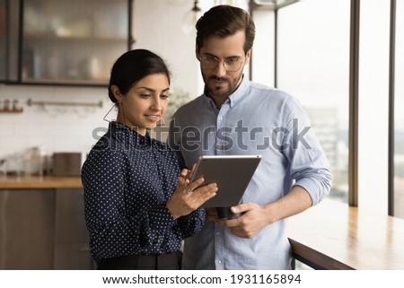 Close up diverse colleagues using tablet together, discussing online project, smiling Indian businesswoman and Caucasian businessman wearing glasses looking at device screen, standing in office Royalty-Free Stock Photo #1931165894