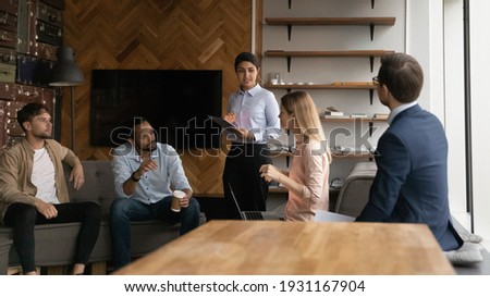 Diverse colleagues discussing project strategy, business partners sharing ideas at corporate meeting in office, confident Indian businessman mentor standing, leading briefing, instructing interns