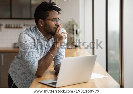 Close up dreamy African American man wearing glasses looking to aside out panoramic window, visualizing, pondering new opportunities or online project strategy, standing at table with laptop Royalty-Free Stock Photo #1931168999