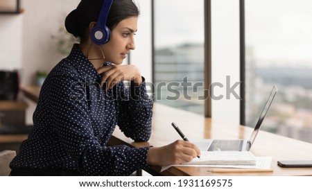 Close up focused Indian woman wearing headphones taking notes, using laptop, intern watching webinar, training, motivated student listening to lecture, sitting at desk near window, studying online