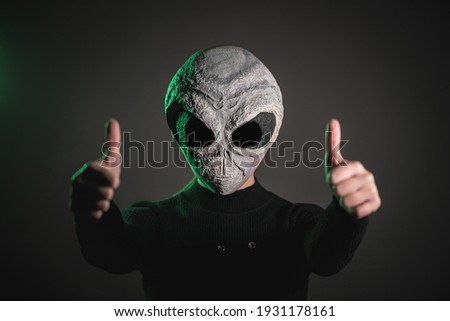An alien is showing a thumbs up gesture on the dark background.