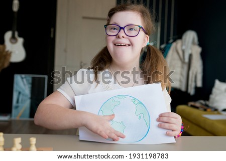 Portrait of smiling teenage girl with Down syndrome holding drawing of the planet Earth at home. Disabled child showing creative artwork. Down syndrome day.