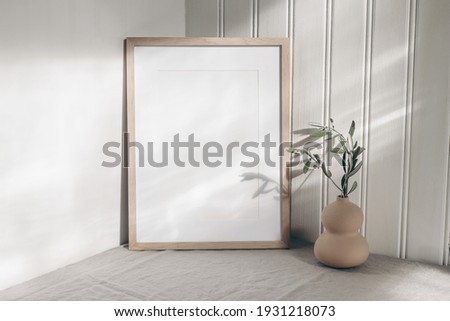Portrait empty wooden frame mockup in sunlight. Olive branch in modern organic shaped vase. Beige linen table cloth. White wainscot wall paneling background. Scandinavian interior, home design. Art.