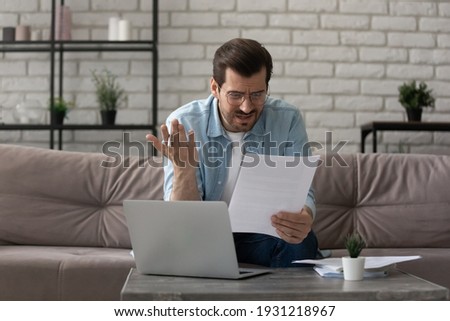 What an outrage. Annoyed young man sit on sofa work with papers at home office read mail letter with bad news unexpected debt. Anxious self employed guy worried by bank report having financial problem Royalty-Free Stock Photo #1931218967