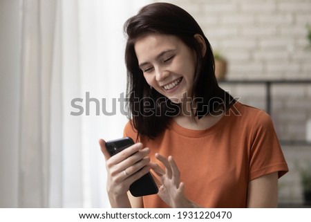 Happy teenage female stand at living room work on smartphone gadget online use wifi internet. Laughing young woman touch screen of modern phone device chat having fun scroll web pages read funny news