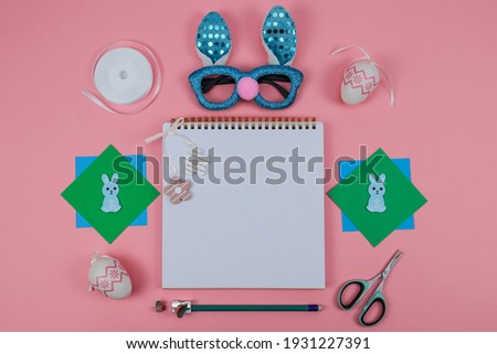 Notepad, scissors, glasses, pencil, colored paper and a bunny on a pink background with a place for text in the middle, close-up top view.