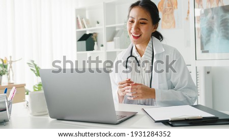 Young Asia lady doctor in white medical uniform with stethoscope using computer laptop talking video conference call with patient at desk in health clinic or hospital. Consulting and therapy concept. Royalty-Free Stock Photo #1931284352