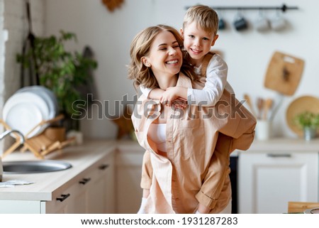 Happy woman giving piggyback ride to cheerful boy while playing in kitchen at home together Royalty-Free Stock Photo #1931287283