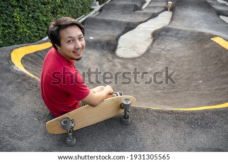 Asian man sitting with surfskate or skate board in pumptrack skate Park when sunrise time over photo blur of pumptrack curve, extream sport, healthy and exercise, fashion in covid19 concept