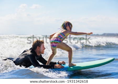 Little surf girl - young surfer learn to ride on surfboard with instructor at surfing school. Active family lifestyle, kids water sport lessons, swimming activity in summer camp. Vacation with child. Royalty-Free Stock Photo #1931342618