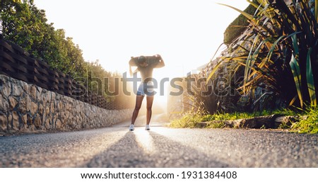 female in trendy wear standing on asphalt road in sunrays holding board for skating on free time in evening, trendy casually dressed woman enjoying active hobby and lifestyle outdoors