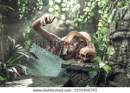 Funny tourist exploring the forest and ancient ruins, she is taking a selfie with a human skull using her smartphone