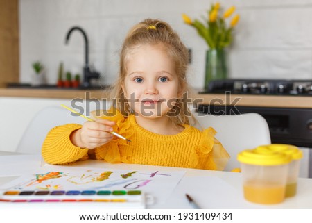 Cute little girl in yellow sweater painting at home. Portrait of 3 years old smiling kid with brush in hands. Concept of early childhood education, hobby, talent, preschool leisure and parenting