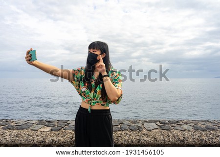 Asian girl taking a selfie. She is posing. In the background you can see the sea. She wears a floral shirt and a mask.