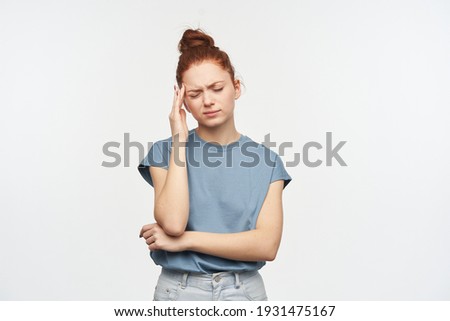 Stressed looking woman, girl with ginger hair gathered in a bun. Wearing blue t-shirt and jeans. Massaging her temple, suffer from migraine. Stand exhausted, isolated over white background