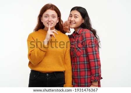 People and lifestyle concept. Two hipster friends. Girl whisper to her friend, showing silence sign. Wearing yellow sweater and checkered shirt. Watching at the camera, isolated over white background