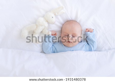 baby boy sleeping on the bed lying on his back with a stuffed toy hares in blue pajamas hands up, healthy newborn sleep Royalty-Free Stock Photo #1931480378