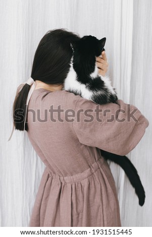 Stylish woman in linen dress hugging cute cat on background of pastel fabric. Simple slow living. Young female in boho rustic dress holding adorable black and white kitty, lovely moment