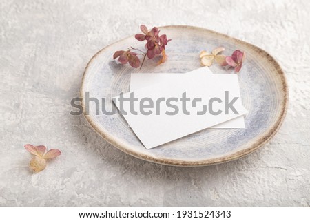 White paper invitation card, mockup with dried hydrangea flowers on ceramic plate and gray concrete background. Blank, side view, still life, copy space.