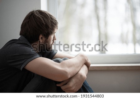 Sad young man sitting on the floor looking through the window Royalty-Free Stock Photo #1931544803