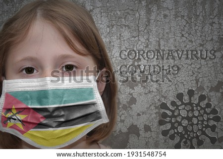 Little girl in medical mask with flag of mozambique stands near the old vintage wall with text coronavirus, covid, and virus picture. Stop virus