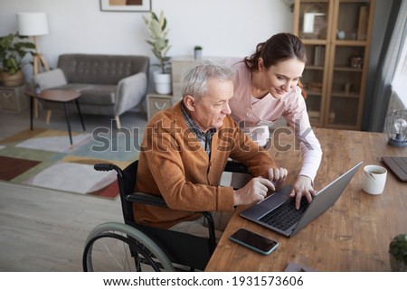 Portrait of senior man in wheelchair using laptop at retirement home with nurse assisting him, copy space Royalty-Free Stock Photo #1931573606