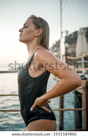 A beautiful young attractive woman in a black swimsuit with a beautiful figure stands under a summer shower on the pier by the sea. She is enjoying her vacation. Selective focus