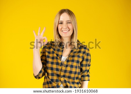 Young woman wearing plaid shirt standing over yellow isolated background smiling positive doing okay sign with hand and fingers. Successful expression.