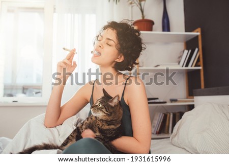 Adult caucasian woman smoking joint with cannabis sitting on the bed with her pets Royalty-Free Stock Photo #1931615996