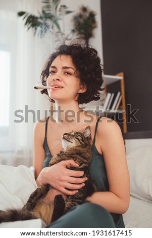 Young woman feeling great with her cat in home while sitting at the bed at the bedroom and smoking. Striped cat looking at his pretty female owner. Stock photo
