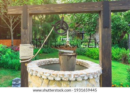 Beautiful artesian well made by stones and wheel pulley with metal bucket and rope in peaceful garden atmosphere. Retro stone water well in rural area. Garden decoration with antique items. Royalty-Free Stock Photo #1931659445