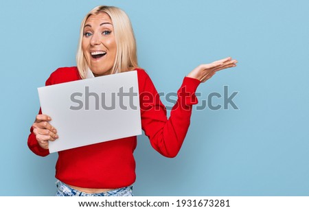 Young blonde woman holding blank empty banner celebrating victory with happy smile and winner expression with raised hands 
