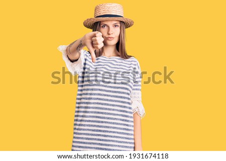 Beautiful caucasian woman wearing summer hat looking unhappy and angry showing rejection and negative with thumbs down gesture. bad expression. 