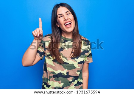 Young beautiful brunette woman wearing camouflage military t-shirt over blue background smiling with an idea or question pointing finger up with happy face, number one