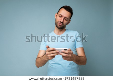 Photo of serious handsome young man with beard wearing everyday blue t-shirt isolated over blue background holding and using mobile phone communication online on the internet looking at gadjet display
