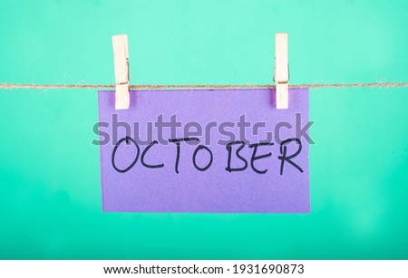 
October word written on a Purple color sticky note hanging with a wire in a Cyan background