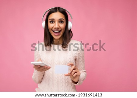 Photo of beautiful young smiling brunette woman wearing pink casual sweater isolated over pink background wall wearing white bluetooth wireless headphones and listening to music and using mobile phone