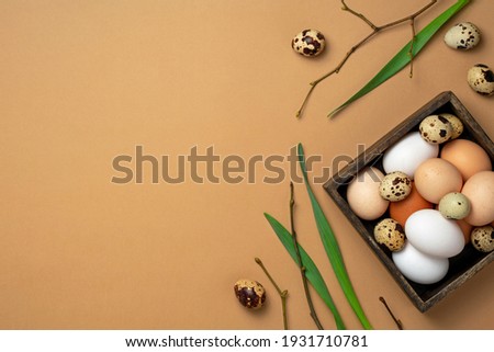 Chicken and quail eggs in wooden box with branches and green leaves on beige background. Easter composition. Top view, copy space, flat lay.