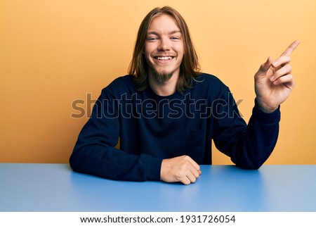 Handsome caucasian man with long hair wearing casual clothes sitting on the table with a big smile on face, pointing with hand and finger to the side looking at the camera. 