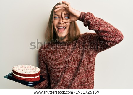 Handsome caucasian man with long hair holding carrot cake smiling happy doing ok sign with hand on eye looking through fingers 