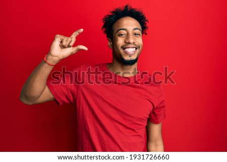 Young african american man with beard wearing casual red t shirt smiling and confident gesturing with hand doing small size sign with fingers looking and the camera. measure concept. 