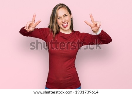 Young blonde woman wearing casual clothes smiling with tongue out showing fingers of both hands doing victory sign. number two. 