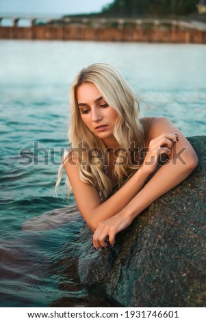 Young blonde woman wear white bikini and sit near stone. sunset beach.
