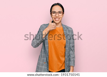 Young hispanic girl wearing business jacket and glasses doing happy thumbs up gesture with hand. approving expression looking at the camera showing success. 